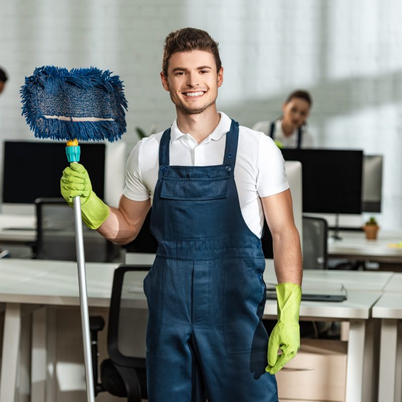 cheerful cleaner holding mop while looking at camera near multicultural colleagues