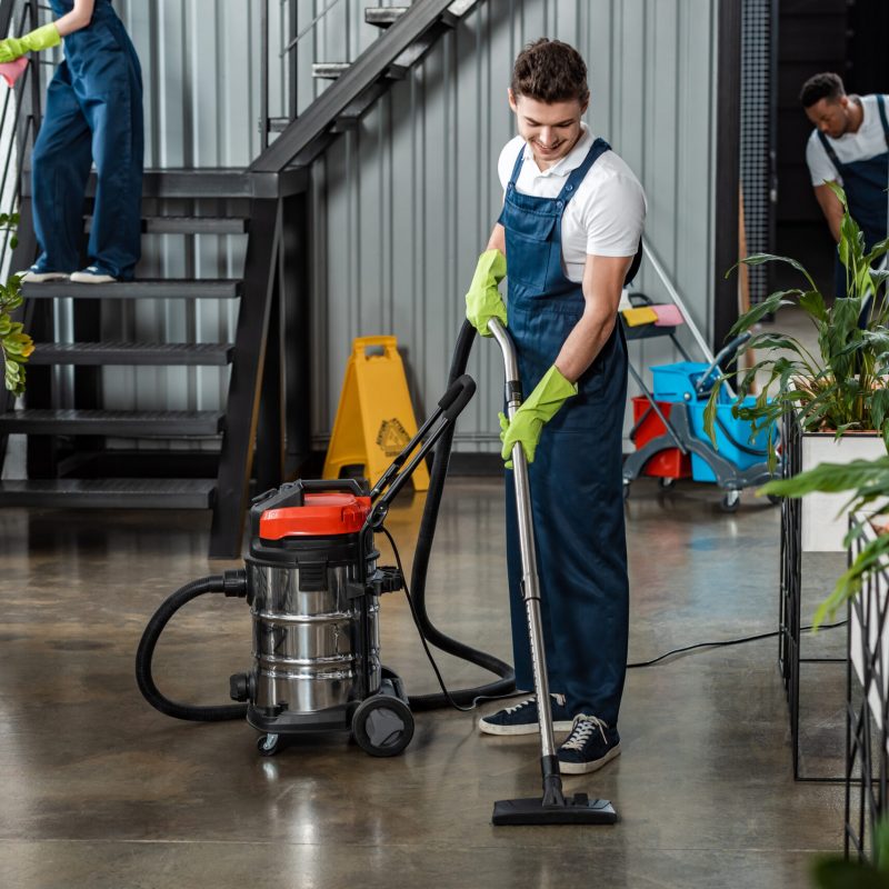 young cleaner cleaning floor with vacuum cleaner near multicultural colleagues